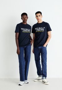 Mørkeblå T-shirts med "RED BULL RACING" grafik i hvid og gul, kombineret med blå jeans og afslappede sneakers, sat mod en lys baggrund.