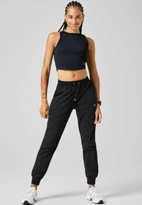 Sort crop-top og sorte joggers med elastiske manchetter, lavet af glat stof. Har justerbar talje og sidelommer. Hvide sneakers.