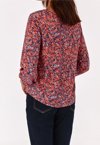Blomstret bluse i livlig rød med blå og gule detaljer, lange ærmer med samlet manchet og rund kant. Sammen med mørke jeans.