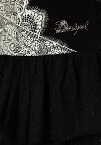 Tissu noir avec motif en dentelle blanche et « Desigual » brodé en fil argenté, montrant des textures superposées et des détails complexes.