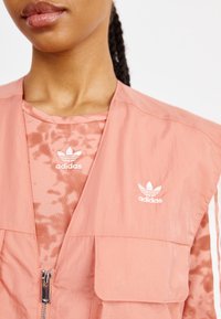 Närbild av en person som bär en rosa Adidas zip-väst över en mönstrad rosa Adidas skjorta, båda med vita Adidas-logotyper.