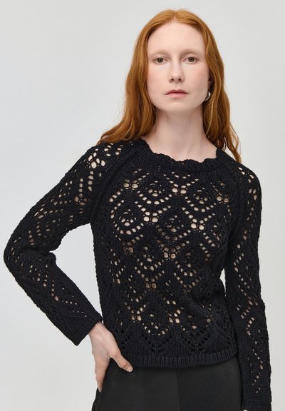 Femme aux longs cheveux roux portant un pull en crochet noir à manches longues avec des motifs géométriques et un col rond, posant avec une main sur la hanche.
