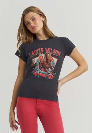 Frau mit langen Haaren trägt ein schwarzes T-Shirt mit einem Pferd, Hufeisen, Rosen, Spielkarten und dem Schriftzug "Lainey Wilson", kombiniert mit roten Hosen.
