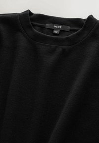 Pull noir à col rond avec une étiquette de marque "NEXT" et une étiquette "Made in China" à l'intérieur du col, présenté sur une surface plane.