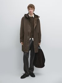 Bruine jas met capuchon over een bruine trui, gecombineerd met grijze broek en zwarte schoenen. Model houdt een donkerbruine suède tas vast.