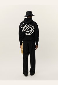 Zwarte hoodie met wit logo, gecombineerd met zwarte broek. Een geel patroonaccessoire hangt aan de zijkant. Achteraanzicht.