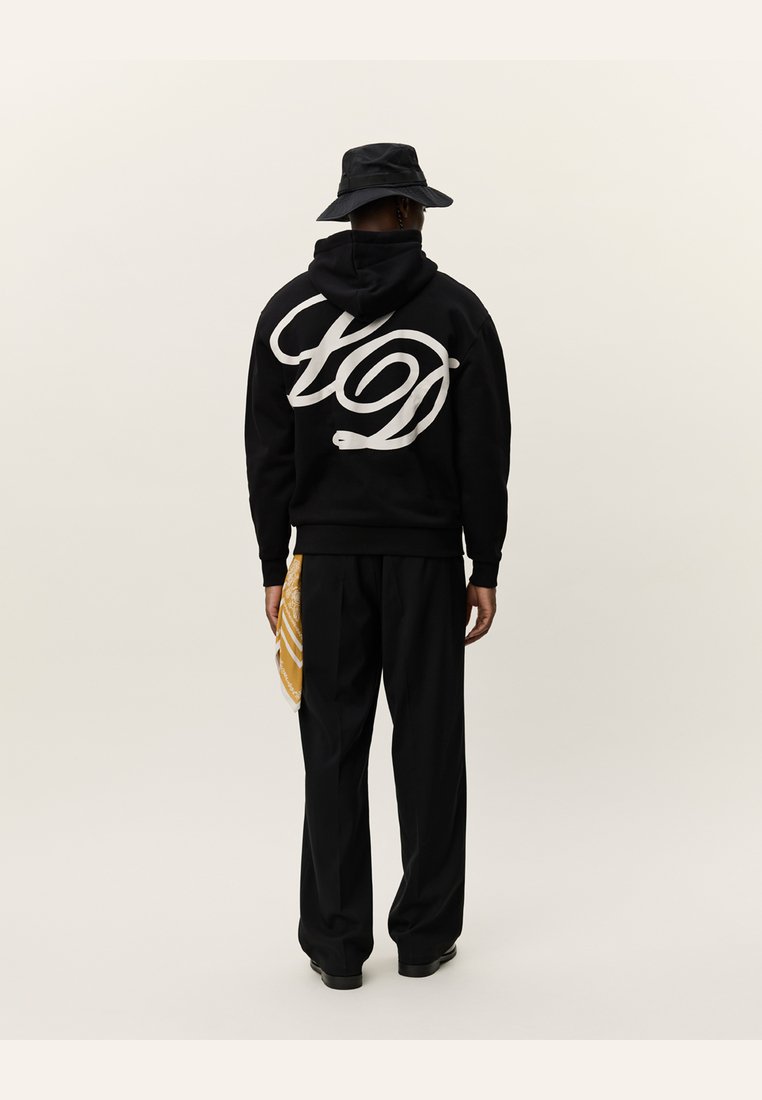 Schwarzer Kapuzenpullover mit weißem Logo, kombiniert mit schwarzen Hosen. Ein gelbes, gemustertes Accessoire hängt von der Seite herab. Rückansicht.