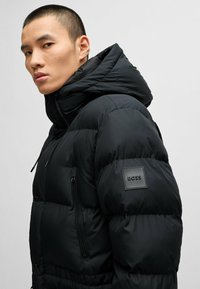 Black puffer jacket with a high collar, zip pocket, and textured logo patch. Features a quilted design and adjustable hood.