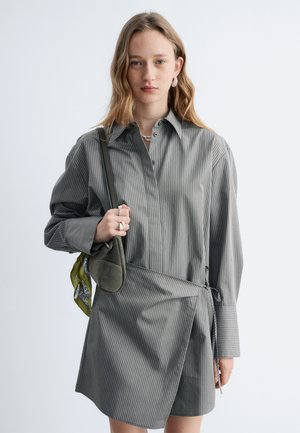 Junge Frau mit langen Haaren trägt ein graues Wickelkleid mit Nadelstreifen, silbernen Schmuck und eine olivgrüne Umhängetasche mit einem gemusterten Schal.