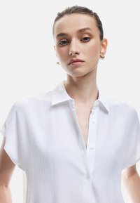 Camicia bianca a maniche corte con bottoni, realizzata in un tessuto testurizzato e dotata di colletto. Presenta una patta con bottoni e una vestibilità ampia.