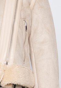 Rukáv béžové semišové bundy s vertikální stříbrnou zipovou kapsou a detaily z krémové ovčí kožešiny.