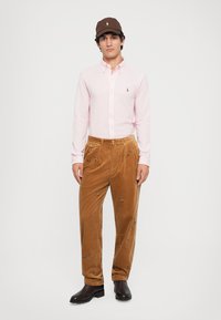 Cămașă roz deschis cu mâneci lungi, pantaloni din bumbac cord din culoare maro cu broderie florală și o șapcă maro. Botine negre până la gleznă completează ținuta.