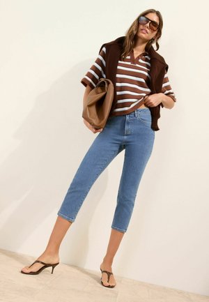 Žena v hnedo-bielom pruhovanom tričku s krátkym rukávom, modrých krátkych džínsoch, hnedom svetri prehodenom cez ramená, slnečných okuliaroch a hnedých sandáloch na podpätku.