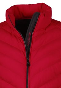 Chaqueta acolchada roja con cuello alto, con cremallera negra y logo "YOU ARE" en el forro interior. Costuras en patrón de chevron por toda la prenda.