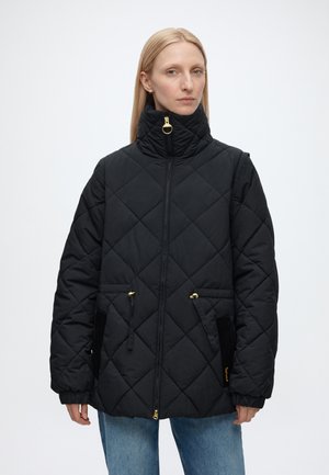 Frau trägt eine schwarze gesteppte Jacke mit hohem Kragen, goldenem Reißverschluss und Kordelzugtaschen, steht vor einem einfarbigen hellen Hintergrund.