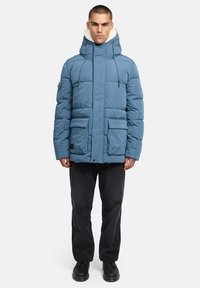 Plava podstavljena parka jakna s visokim ovratnikom i izvlakama, koja ima više prednjih džepova i kapu obloženu mekim materijalom. Crne hlače i cipele.