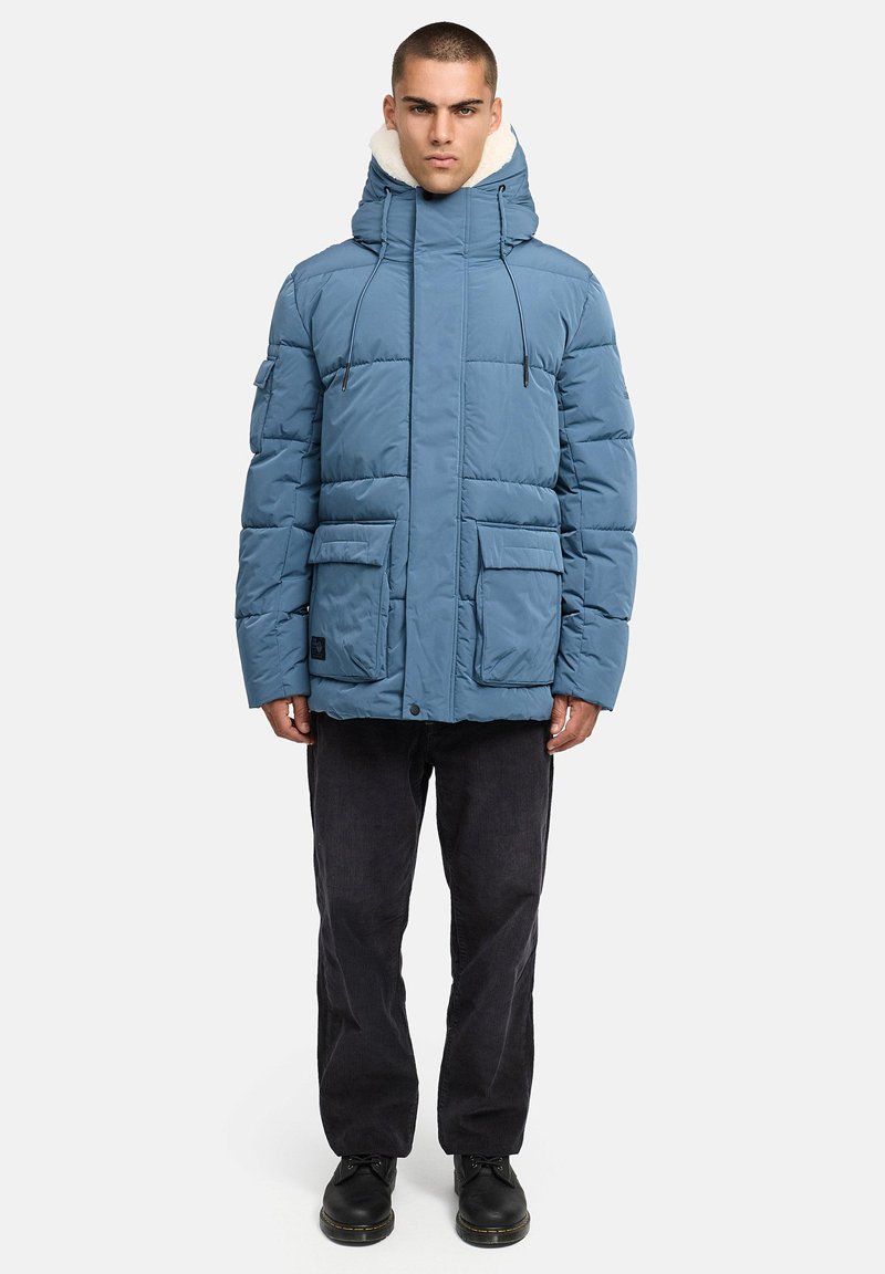 Plava podstavljena parka jakna s visokim ovratnikom i izvlakama, koja ima više prednjih džepova i kapu obloženu mekim materijalom. Crne hlače i cipele.