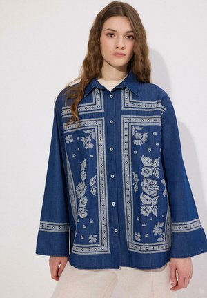 Kobieta o długich brązowych włosach ubrana w oversize'ową niebieską koszulę na guziki z białym kwiatowym haftem oraz beżowe spodnie na białym tle.