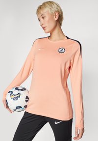 Långärmad persikofärgad sporttröja med marinblå detaljer och Chelsea FC-logotyp, tillsammans med svarta träningsbyxor och håller en mönstrad fotboll.
