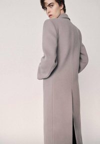 Lang, lysegrå frakke lavet af blødt stof med skræddersyet design, åben front og et slids bagpå for nem bevægelse.