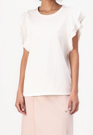 Kobieta ubrana w białą, marszczoną bluzkę z krótkim rękawem, zestawioną z jasno beżową spódnicą z guzikami po boku.