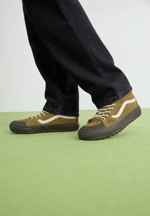 Braune Wildledersneaker mit Gummisohlen, ausgestattet mit cremefarbenen Akzenten und runden Schuhschnürsenkeln, kombiniert mit schwarzen Jeans auf einer grünen Fläche.