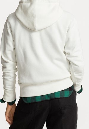 Personne portant un sweat à capuche blanc sur une chemise à carreaux verte et noire avec un pantalon noir, vue de dos.