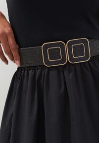 Ceinture tissée noire avec deux boucles carrées aux accents dorés et une finition texturée, portée sur une jupe noire à tissu froncé.