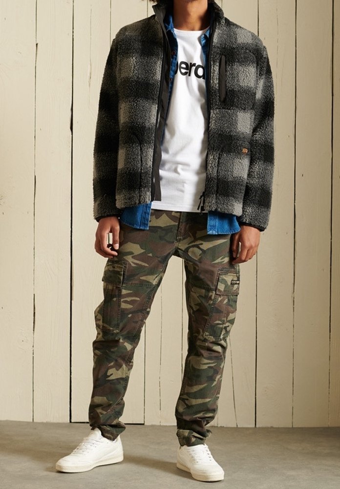 Personne portant une veste polaire à carreaux grise et noire, un t-shirt blanc imprimé, une chemise bleue, un pantalon cargo camouflage et des baskets blanches, debout à l'intérieur.