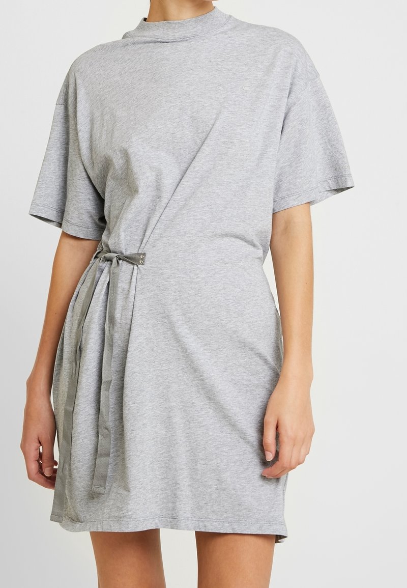Femme portant une robe gris clair à manches courtes, avec un col montant et une ceinture en tissu nouée sur le côté gauche.
