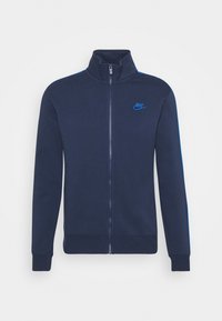 Marinblå zip-up jacka med hög krage, med blå randiga detaljer och broderad logotyp på bröstet, tillverkad av mjukt, texturerat tyg.