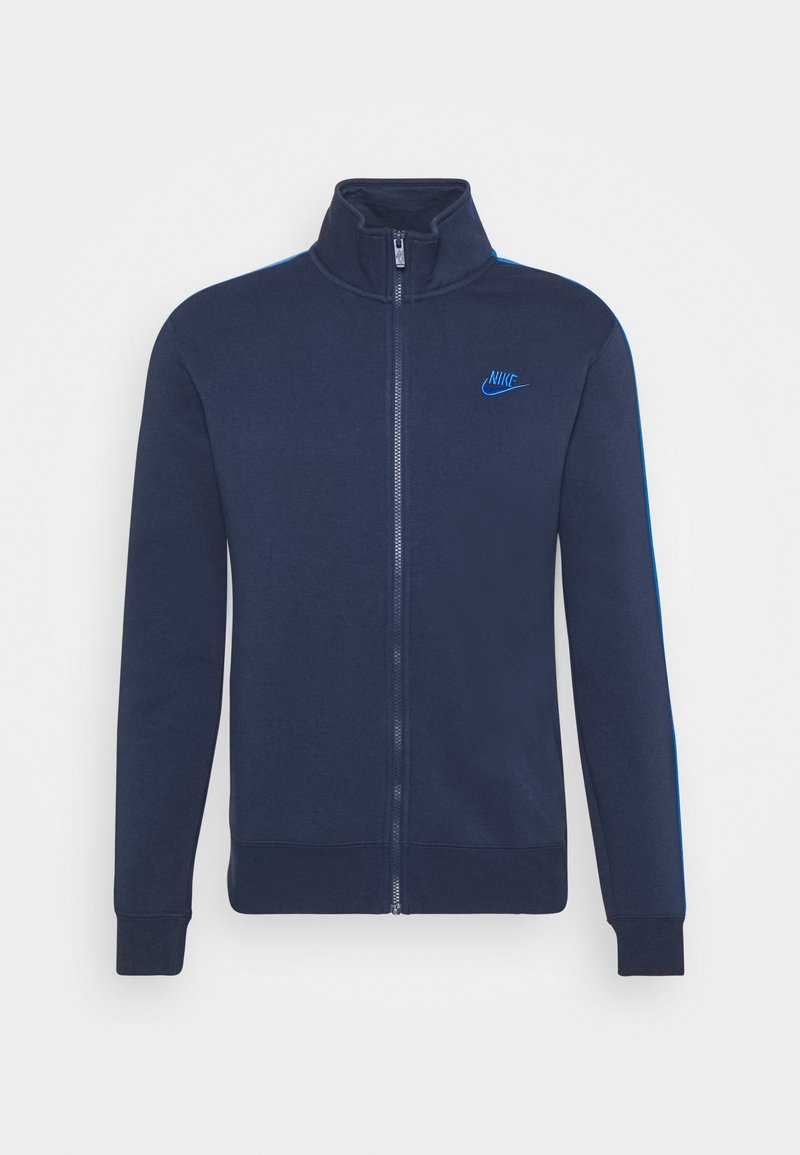 Marinblå zip-up jacka med hög krage, med blå randiga detaljer och broderad logotyp på bröstet, tillverkad av mjukt, texturerat tyg.