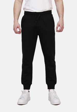 Uomo con pantaloni jogger neri con polsini elastici e sneakers bianche, in piedi davanti a uno sfondo semplice.