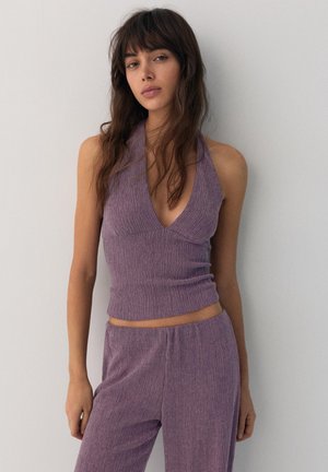 Femme aux longs cheveux bruns portant un haut sans manches violet texturé et un pantalon assorti, debout contre un mur blanc uni.