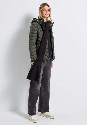 Groene pufferjack met capuchon en ritszakken, gecombineerd met zwarte jeans en een lange zwarte sjaal. Witte en beige sneakers maken de look compleet.