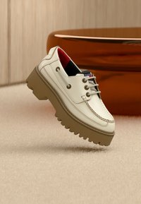 Άσπρο δερμάτινο παπούτσι τύπου boat με χοντρή σόλα, μπεζ κορδόνια και κόκκινη επένδυση στο εσωτερικό, τοποθετημένο σε ανοιχτό χαλί με καφέ έπιπλα στο φόντο.