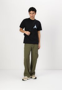 Black cotton t-shirt with a blue graphic design, paired with loose olive cargo pants and beige sneakers, standing against a white background.