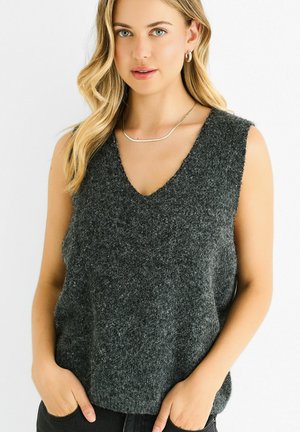 Gray wool-knit sleeveless sweater with a deep V-neck, soft texture, and relaxed fit. Model poses with hands in pockets, wearing a simple necklace.