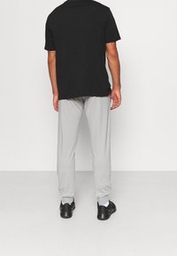 En man som står vänd mot en slät vit vägg, klädd i en svart t-shirt, ljusgrå joggers, vita strumpor och svarta sneakers.