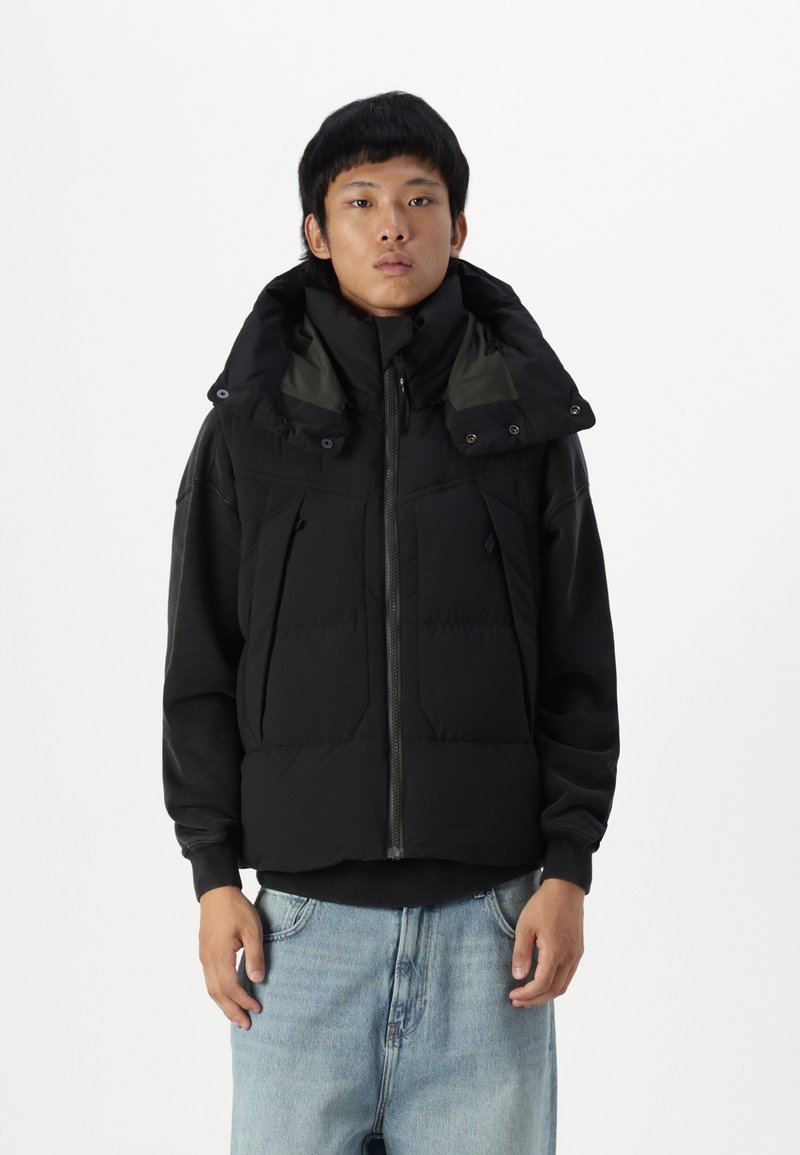 Chaleco de plumas negro con cuello alto, cierre de cremallera y múltiples bolsillos frontales. La capa interior tiene un forro contrastante de color verde oscuro.