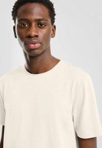 Lichtbeige t-shirt met ronde hals, korte mouwen en een gladde textuur, gedragen door een jonge man tegen een neutrale achtergrond.