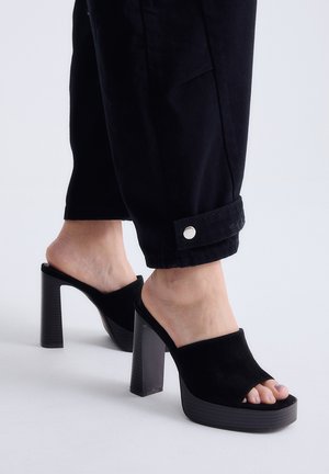 Pies que llevan mules de plataforma de tacón alto en negro, combinados con pantalones negros que tienen puños con botones a presión en los tobillos.