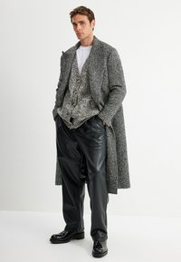 Cappotto grigio a spina di pesce sopra un gilet grigio a fantasia, abbinato a pantaloni neri plissettati e scarpe nere lucide. Sotto indossa una semplice t-shirt bianca.