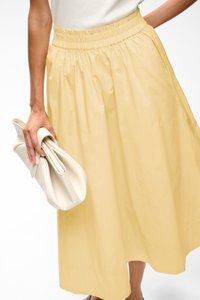 Personne portant une jupe midi jaune clair et un haut blanc, tenant une pochette blanche douce dans une main sur un fond neutre.