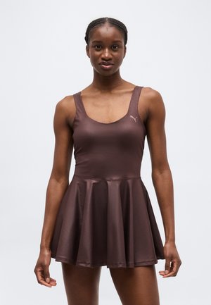 Žena s vrkočmi, oblečená v bezruksákových hnedých športových šatách s rozšírenou sukňou a logom Puma na hrudi, stojaca na svetlom pozadí.