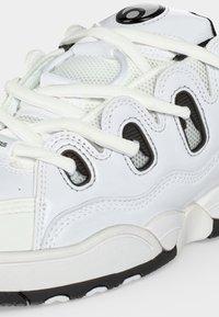 Gros plan d'une basket blanche avec des panneaux en mesh, plusieurs œillets, des lacets blancs, des accents noirs, et une semelle épaisse noire et blanche.