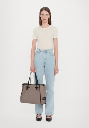 Tote bag van beige stof met zwarte accenten en handvatten, met een gestructureerde vorm en trekkoordsluiting. Model draagt een lichte top en jeans.