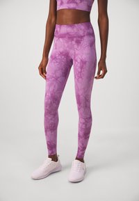 Fialové legíny s tie-dye vzorom majú vysoký pás a pružnú tkaninu. Doplnia ich biele športové tenisky so štruktúrovaným povrchom.