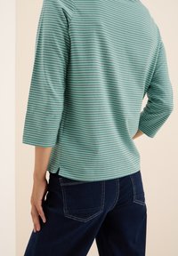 Personne portant un t-shirt rayé teal et blanc à manches longues et un jean bleu foncé, vu de dos, avec une main sur la hanche.