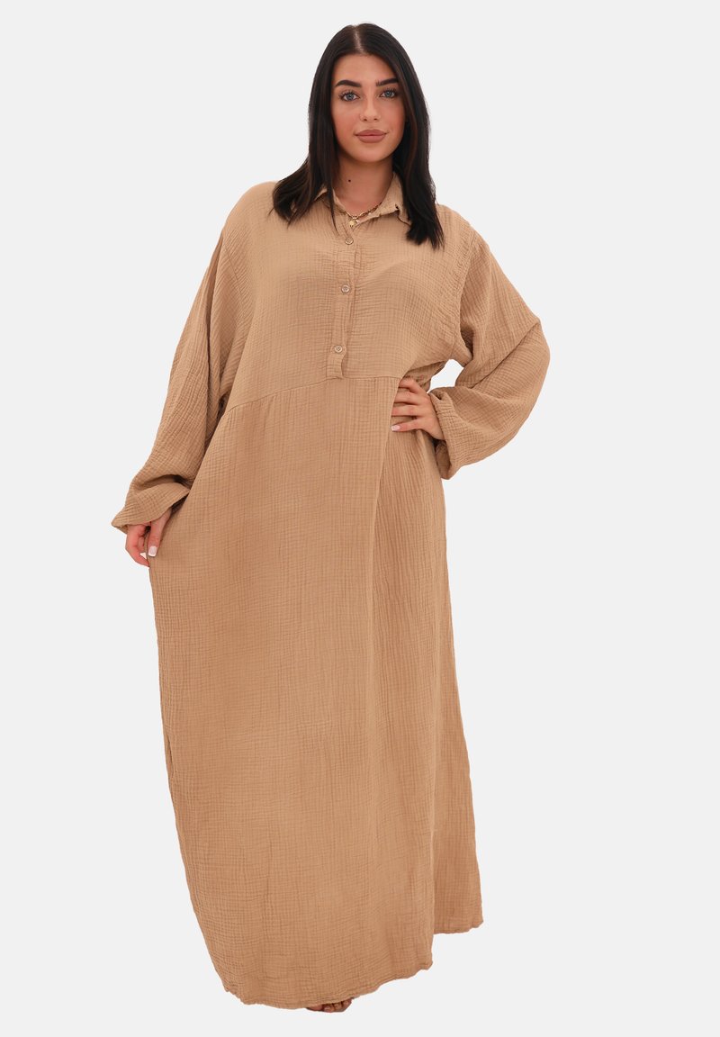 En kvinde står selvsikkert i en lang, beige knap-til-kjole med oversized ærmer, hvilket fremhæver en afslappet og modebevidst stil.